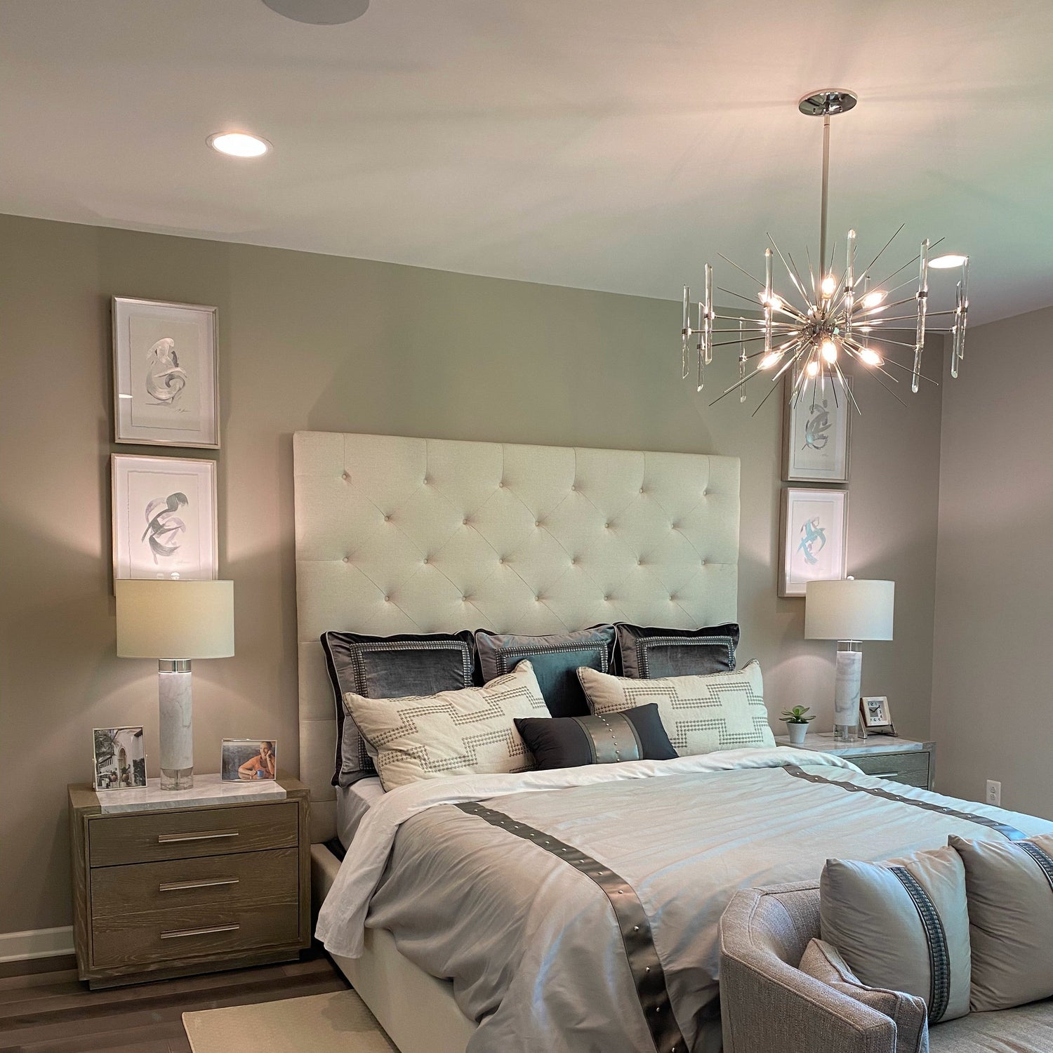 Image of a bedroom with a decorative chandelier, ceiling recessed lighting, and table lamps illuminating the space well. The room is a beige tone, with plenty of decorations on the walls and other surfaces.